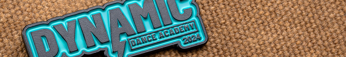 Close-up of a teal and dark grey Dynamic Dance Academy 2024 pin with a lightning bolt through the wordmark