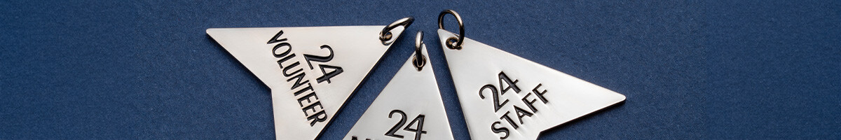 Three polished silver arrow-shaped charms engraved 24 Volunteer, 24 and 24 Staff on a navy background