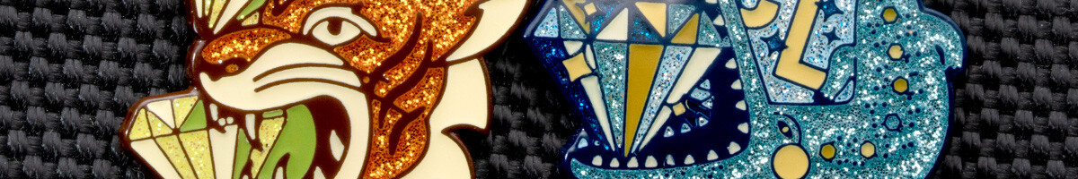 Close-up of two glitter enamel pins, an orange tiger with cut-gem facets and a blue design with a faceted diamond, showing the flake suspended in the cured enamel