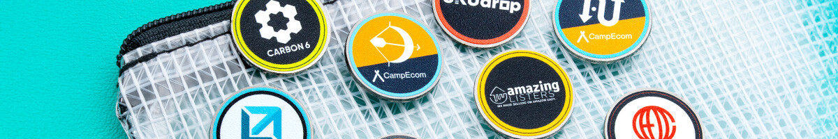 A dozen round printed company logo pins arranged on a mesh travel pouch, including Carbon 6, CampEcom, SKUdrop and Amazing Listers designs