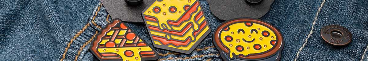 Three pizza-themed soft enamel pins — a pizza slice, a bacon and cheese wedge, and a smiling pizza — pinned to a denim jacket